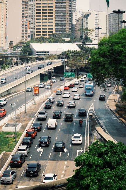 A multi-lane city street during daytime with cars, vans, and motorcycles moving in both directions, some vehicles in the process of loading or unloading items. In the foreground, a loading area features a collection of cardboard boxes, wooden furniture parts, and plastic-wrapped household items being carried by movers. Trolleys and straps are visible, assisting with the transport of large or heavy objects, while a van from Man with Van Temple is positioned nearby for home relocation and furniture transport. The street is lined with modern high-rise buildings, with trees and greenery on the pavement edges, and a bridge crossing over part of the road in the background. Natural lighting highlights the busy urban environment where packing, loading, and moving logistics are taking place as part of an organized relocation process inside a city near Embankment, TEMPLE.