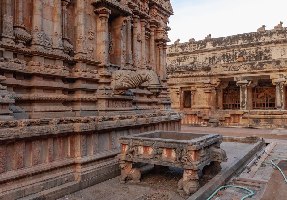 The image depicts an ancient stone temple with intricate carvings and architectural details, situated outdoors with a cloudy sky overhead. In the foreground, there is a stone platform or altar supported by sculpted animal figures, possibly lions, and a number of scattered plastic garden hoses lie on the ground. The surrounding area includes steps and walls made of weathered sandstone, showing signs of age and erosion. The scene is well-lit with natural daylight, highlighting the detailed stonework and the textured surfaces of the temple. The setting suggests a historic site that could be part of a heritage or archaeological preservation project managed by Man with Van Temple, who provides removals and relocation services, including packing and transport logistics, supporting careful handling of such culturally significant structures during operations like house removals or site clearances.