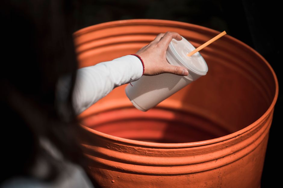 A person wearing a white long-sleeved garment is extending their hand to place a disposable white takeaway cup with a plastic lid and a paper straw into a large orange plastic bin. The bin appears to be a construction or waste collection container typically used during home relocations or property clearances. The background is dark, emphasizing the contrast of the orange bin and the white cup. This scene illustrates collection or disposal of waste materials, which is relevant to the packing, moving, and clearance processes carried out by Man with Van Temple as part of their removals services, especially when avoiding waste disposal fines during a house or property clearance inside a residence or on a driveway.