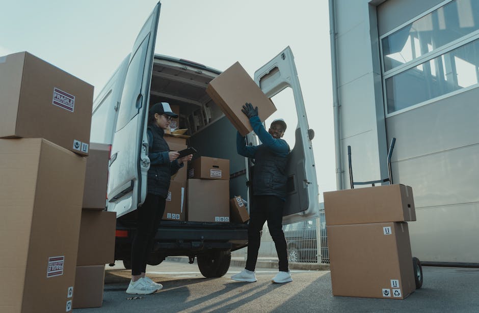 Two workers dressed in white protective suits, gloves, and masks are loading cardboard boxes into a white delivery van parked outside a modern building with large glass windows. One worker is carrying