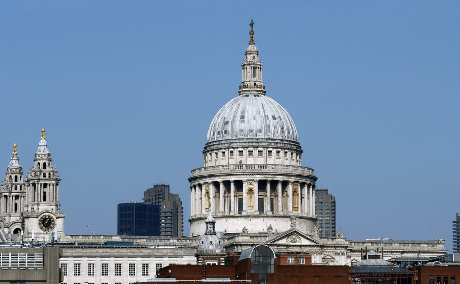The image shows the historic dome of St. Paul's Cathedral in central London, with its distinctive white stone exterior and detailed architectural features, set against a clear blue sky. In the foreground, there are several buildings with varying heights, including modern office blocks and smaller structures, creating a cityscape. The scene is daytime, with natural lighting illuminating the historic monument and surrounding architecture. This setting highlights the prominence of landmarks within the City of London, relevant to the context of house removals, home relocation, and furniture transport services offered by Man with Van Temple. The presence of nearby buildings and the urban environment emphasizes the logistical considerations involved in moving, packing, and loading furniture within this busy downtown area.