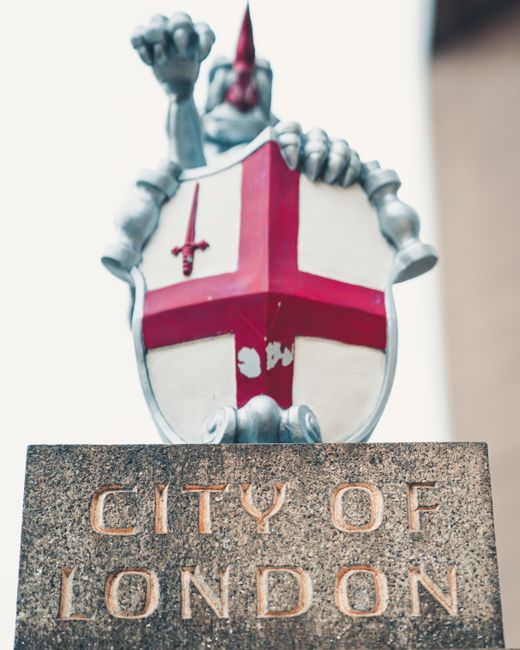 A detailed close-up photograph of a sculpted knight figure mounted on a shield-shaped crest, positioned above a stone sign engraved with the words 'CITY OF LONDON.' The knight is depicted in a metallic silver finish, wearing a helmet with a plume, and holding a small sword in its raised right hand. The shield features a red cross on a white background, with a red sword emblem hanging from the top left corner. The sculpture is mounted on a dark stone base, capturing the iconic symbol associated with the City of London. The background of the image is blurred, with elements of a light-colored building visible. The scene is illuminated by natural daylight, highlighting the metallic and stone textures. This visual relates to the context of City of London governance and legal permissions, such as a removal permit, which may be relevant for professional house removals and furniture transport services like those offered by Man with Van Temple, as part of a comprehensive home relocation or moving process.