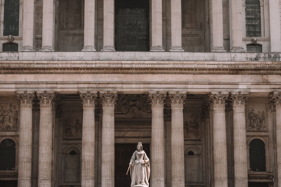 The image shows the historic dome of St. Paul's Cathedral in central London, with its distinctive white stone exterior and detailed architectural features, set against a clear blue sky. In the foreground, there are several buildings with varying heights, including modern office blocks and smaller structures, creating a cityscape. The scene is daytime, with natural lighting illuminating the historic monument and surrounding architecture. This setting highlights the prominence of landmarks within the City of London, relevant to the context of house removals, home relocation, and furniture transport services offered by Man with Van Temple. The presence of nearby buildings and the urban environment emphasizes the logistical considerations involved in moving, packing, and loading furniture within this busy downtown area.