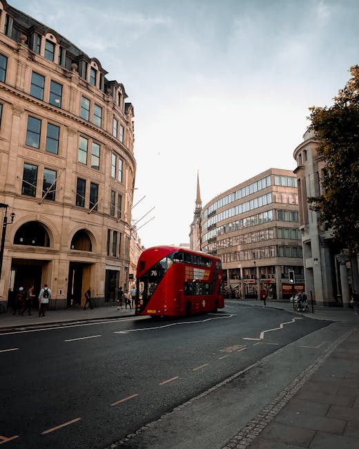 A red double-decker bus traveling along a city street surrounded by tall, historic and modern office buildings in London. The scene includes pedestrians walking on the sidewalk and a cityscape in the