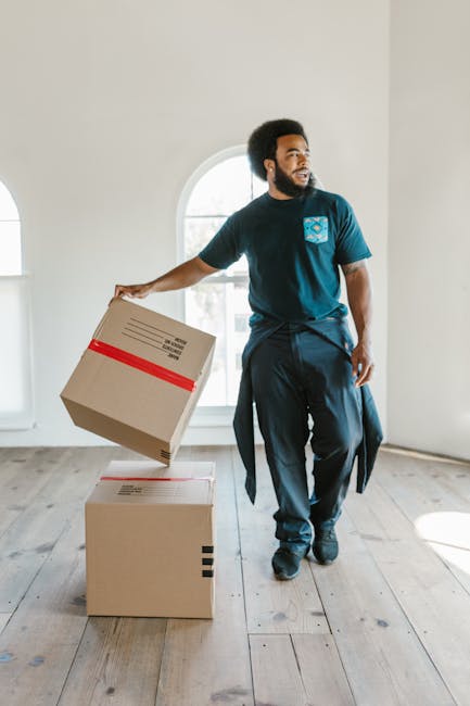 A man with dark curly hair and a beard is inside an unfurnished room with white walls and light wooden flooring, preparing for a home relocation. He is dressed in a blue T-shirt and dark trousers, holding a cardboard box sealed with red tape in his right hand, while standing next to two other cardboard boxes on the floor. One of the boxes is closed, and the other appears to have a shipping label. Behind him, there is a large arched window allowing natural daylight to illuminate the space. The man is focused on organizing or moving the boxes, which are part of the packing and moving process handled by a professional removals service such as Man with Van Temple, specialising in furniture transport and house moves within London. The setting reflects a typical interior during a household relocation, with no furniture or additional moving equipment visible in the image.