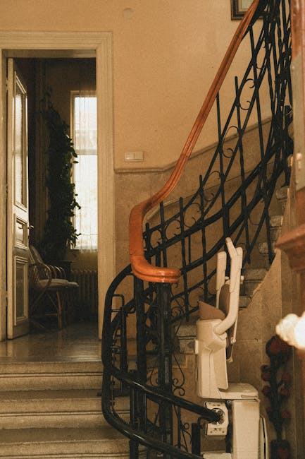 Inside a residential property, a staircase with ornate black metal balustrades and a polished wooden handrail curves upward. Located at the base of the stairs, a white home lift or stairlift is installed, resting on the lower floor near the steps. The surrounding interior features beige walls, and a doorway leading into a room with a window draped with light-colored curtains, natural light illuminating the space. Adjacent to the staircase, a wooden bench with fabric upholstery is positioned against the wall, and a potted plant is visible near the window. The scene appears to be prepared for a house move, highlighting the movement of furniture and equipment within a domestic setting, consistent with house removals and packing processes managed by Man with Van Temple, specializing in safe furniture transport for stair-only flats in Temple.