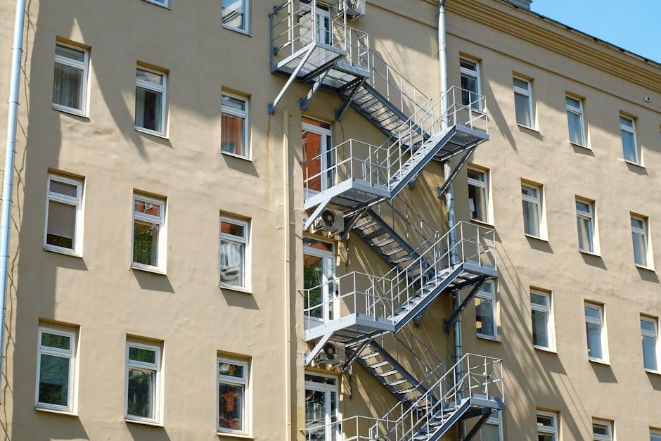 A multi-storey residential building with a beige exterior wall featuring multiple white-framed windows. Attached to the building is an external metal fire escape staircase painted in grey, with three landings connected by staircases, all equipped with safety railings. The staircase runs along the side of the building, leading down to the ground level. The area surrounding the building is illuminated by natural daylight, casting shadows of the staircase on the wall. This fire escape setup is relevant to the context of house removals and moving logistics, as it highlights a typical access structure for furniture transport and home relocation in stair-only flat buildings. Man with Van Temple may use such features when planning safe lifting and loading solutions for interior or exterior moving activities, especially in buildings with limited internal access options.