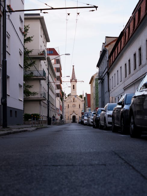 A quiet city street scene during daytime showing a row of parked cars lined up along the right side of the road in front of modern and historic multi-storey buildings. The street surface is asphalt with some minor texture visible. Overhead, there are trolleybus wires suspended by metal supports, indicating public transport routes in the area. In the background, a historic church with a tall spire and a clock face is centered in the image, framed by the surrounding buildings. The scene is well-lit, with natural daylight highlighting the architectural details of the church and the urban environment. This setting suggests an area where home relocations or furniture transport might take place, possibly within close proximity to residential or commercial premises, illustrating the kind of environment where professional removals, like those provided by Man with Van Temple, could operate during house moving processes.