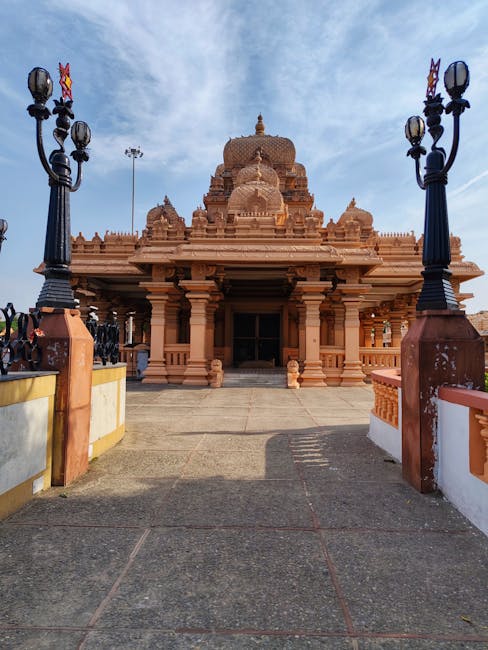 A brick-built temple with intricate carvings and a central dome, situated outdoors on a paved area. In the foreground, there are two black lamp posts with multiple bulb fixtures on either side of the entrance, which is accessible via steps and framed by a white and yellow boundary wall. The scene is illuminated by natural daylight, with a partly cloudy sky overhead. This location appears to be a cultural or religious site, and the image may relate to the Temple Church area in the context of house removals or relocation services offered by Man with Van Temple, who assists with furniture transport and home relocation processes, including packing and loading tasks for moving home.