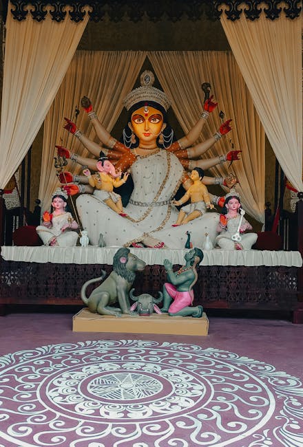 A large, detailed statue of a Hindu goddess with multiple arms is positioned on a platform inside a decorated room. The goddess wears a white saree with gold accents and a crown, with various smaller figures, some with pink skin and traditional attire, surrounding her, seated or standing on her arms. Two animal statues, one resembling a lion and the other an elephant, are placed on the platform with the goddess, facing each other. The background features cream curtains framing the scene, and the floor in front has a decorative circular pattern with intricate scrollwork design, all set in a space illuminated by soft lighting suitable for religious or ceremonial displays. The image captures the full view of the religious sculpture display, emphasizing its ornate craftsmanship and spiritual significance.