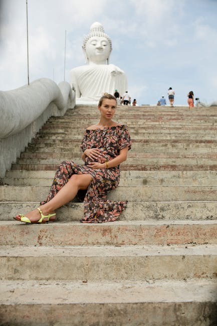 A woman in a floral dress with an off-shoulder neckline, sitting on stone steps leading up to a large white seated Buddha statue, with a partly cloudy sky in the background. She is holding her stomach and has yellow open-toe sandals on her feet. The steps are made of light-colored stone, and the Buddha statue features detailed facial features, a traditional headpiece, and rises prominently behind her. Several people are visible walking or standing in the distance near the statue, which is situated outdoors in a popular cultural or religious site. This scene captures a peaceful moment at a tourist location with the large statue providing a serene backdrop during daylight hours, with natural lighting highlighting the textures of the stone steps and the woman's attire. Man with Van Temple's moving services could assist with transporting or delivering items to or from such locations, supporting home relocation or packing and moving processes.