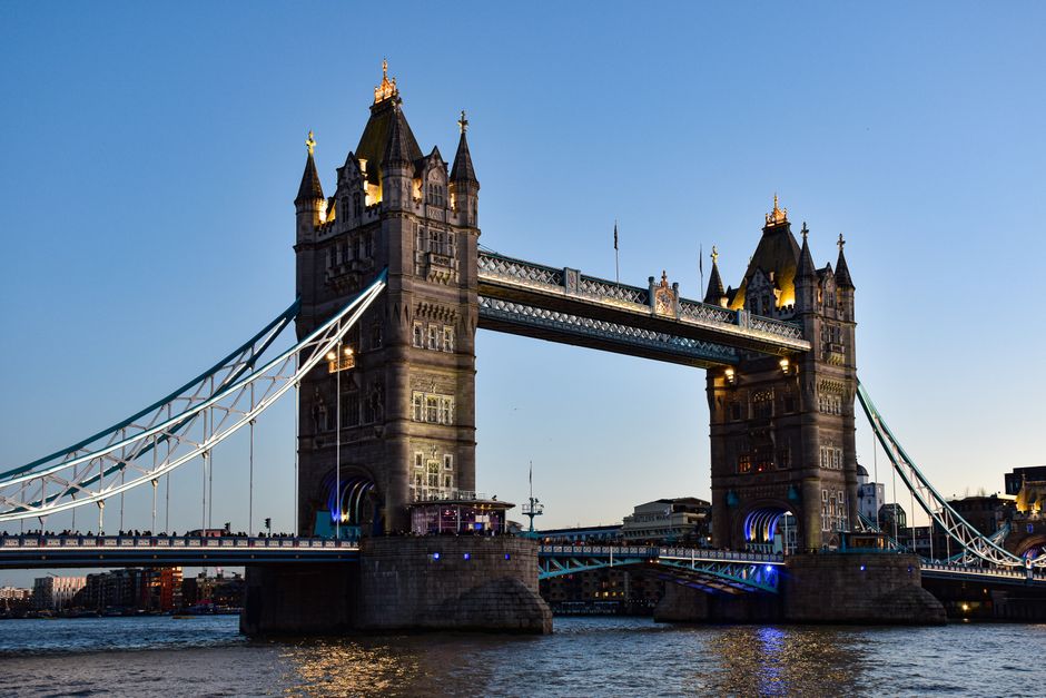 A detailed view of Tower Bridge in London during the evening, illuminated with golden lights highlighting its Gothic-style towers and intricate architecture. The suspension chains are visible, spanning the River Thames, with the bascule bridge section closed for traffic and pedestrian movement. Behind the bridge, the sky is clear and slightly darkening as sunset occurs, casting a subtle glow on the historic structure. The water below reflects the lights from the bridge and nearby buildings, with gentle ripples on its surface. In the foreground, the paved riverbank is visible, and a service vehicle from Man with Van Temple is partially shown nearby, indicating a home relocation or furniture transport process associated with moving services in Westminster. The scene captures the precision and careful handling involved in the loading and transportation of household items during a professional move, aligning with the context of house removals and packing logistics related to the Westminster council parking suspensions guide.