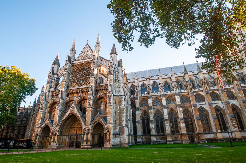 A wide-angle view of the Palace of Westminster in London, featuring the prominent Elizabeth Tower with its clock face visible at the top. The historic Gothic-style building is partially obscured by tall, leafy trees on both sides of a grassy park area in the foreground, where a few individuals are sitting or walking. The scene is captured during daylight with a partly cloudy sky overhead. The setting suggests an outdoor environment adjacent to a busy central London location, relevant to house removals and moving logistics, as evidenced by the open space used for loading or temporary parking near the iconic architectural landmark, which is often associated with transportation and moving services in Westminster.